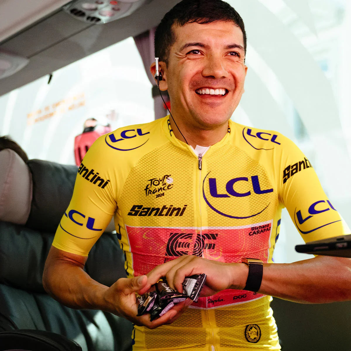 Smiling cyclist in yellow LCL Tour de France 2024 jersey holding TurboChew Bar packs on a bus