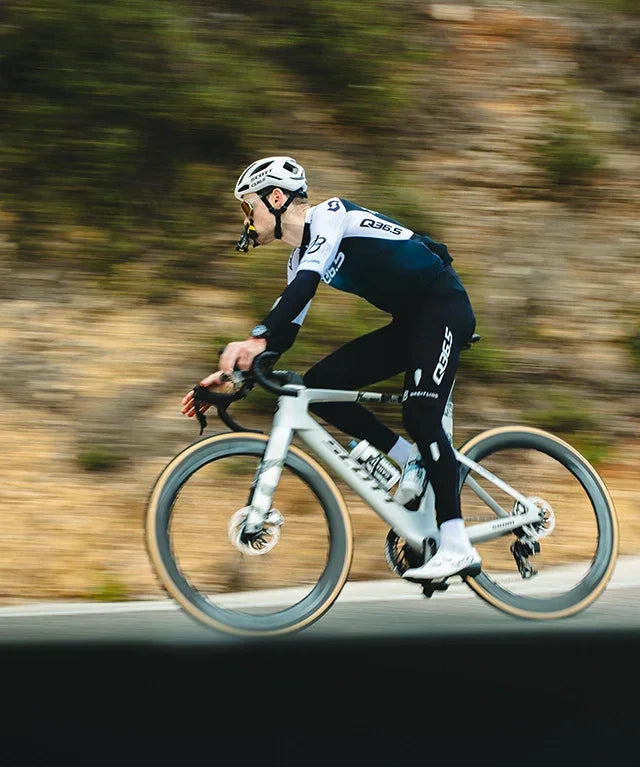 Cyclist in QB365 kit riding white road bike outdoors on a blurred mountain road.