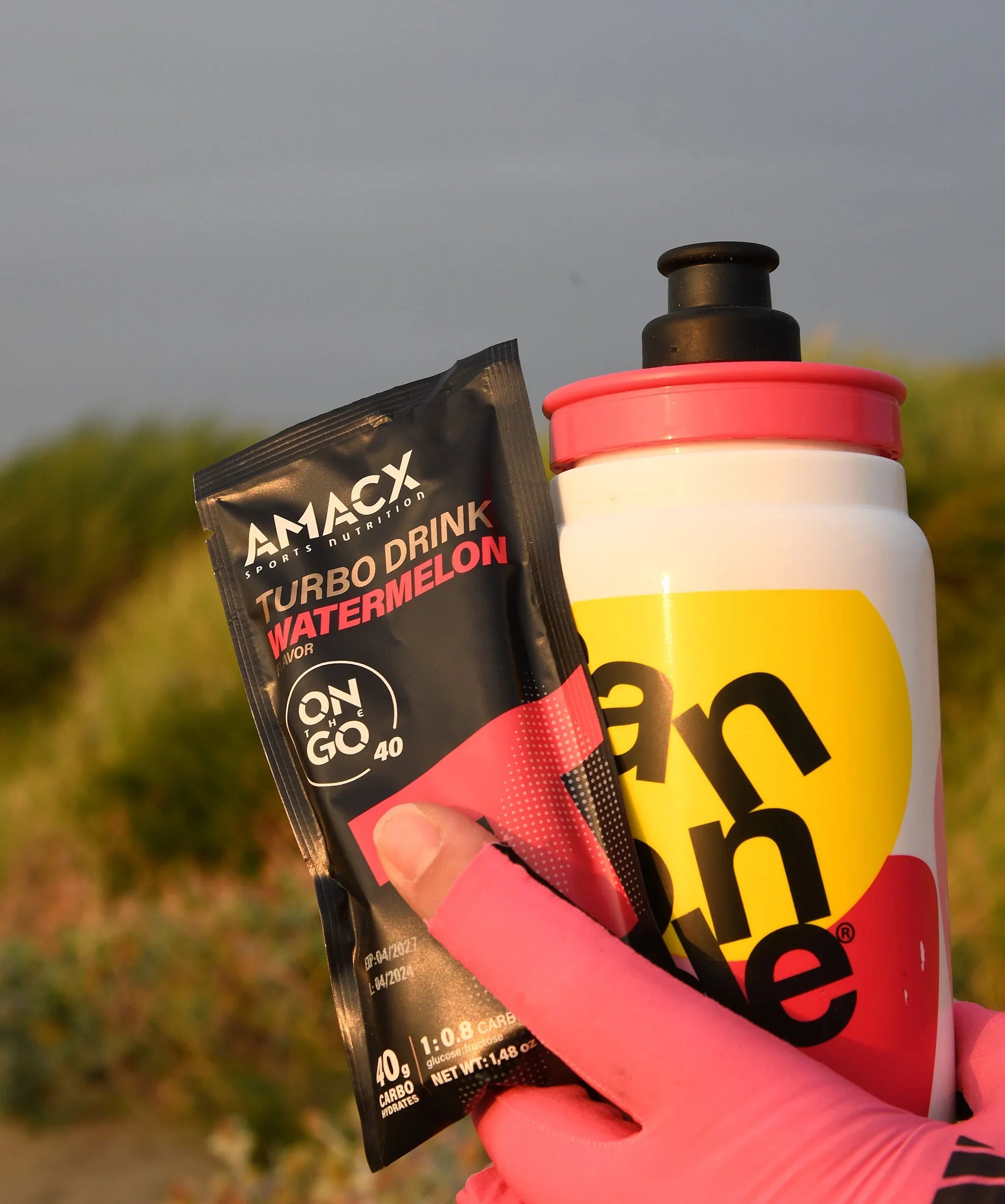 AMACX Turbo Drink Watermelon sachet and yellow sports water bottle held outdoors