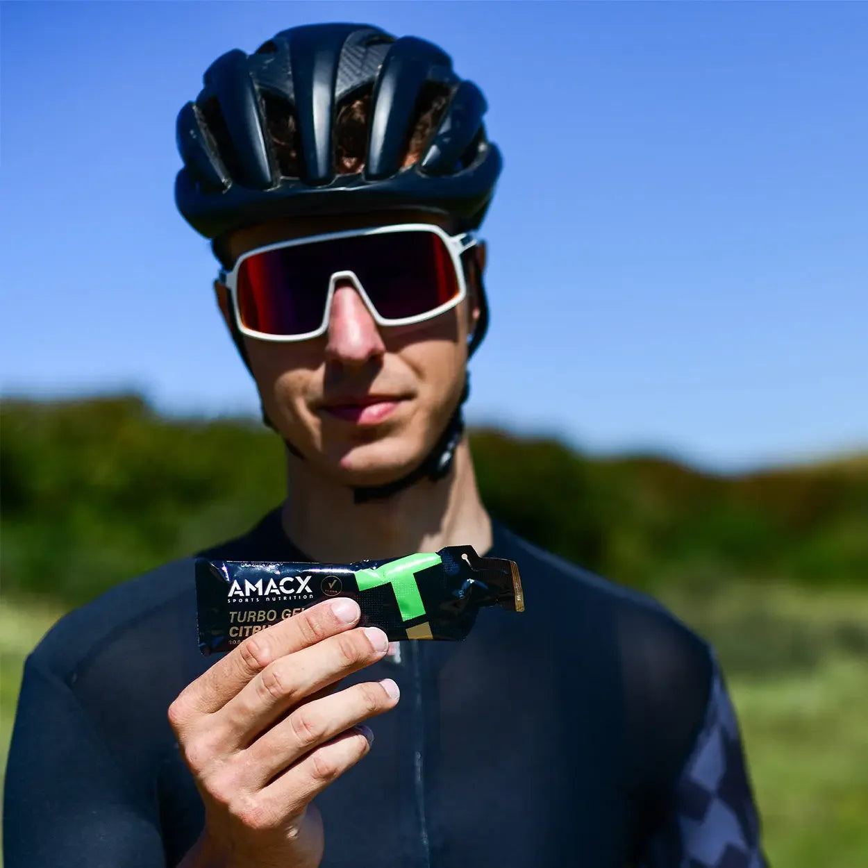 Cyclist wearing helmet and sunglasses holds AMACX Turbo Gel energy gel outdoors