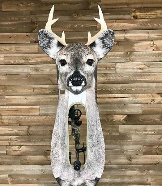 Archery deer target with whitetail antlers on a wooden wall, bow visible behind cutout.