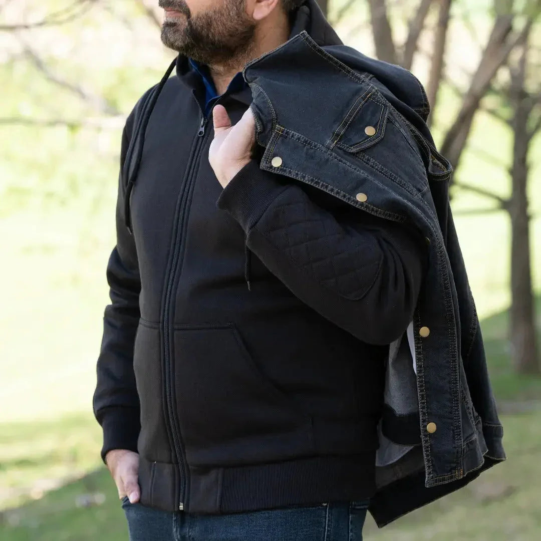 Man in a black zip-up hoodie holding a denim vest outdoors, casual men's fashion