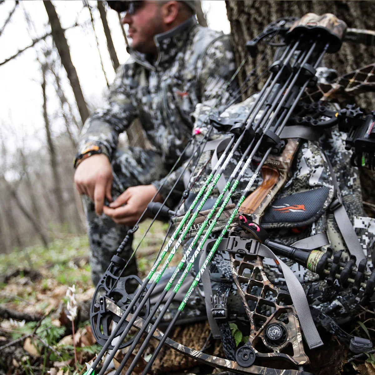 Hunter in camo gear sitting by a tree with a compound bow and arrows in a forest setting