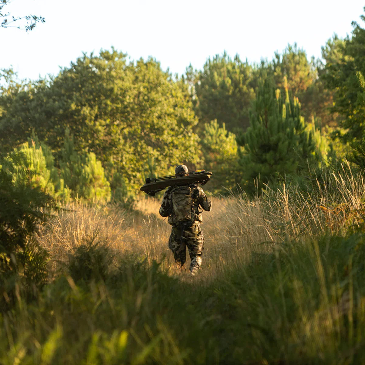 Hunter in camo gear carrying a treestand walking through tall grass in a forest clearing