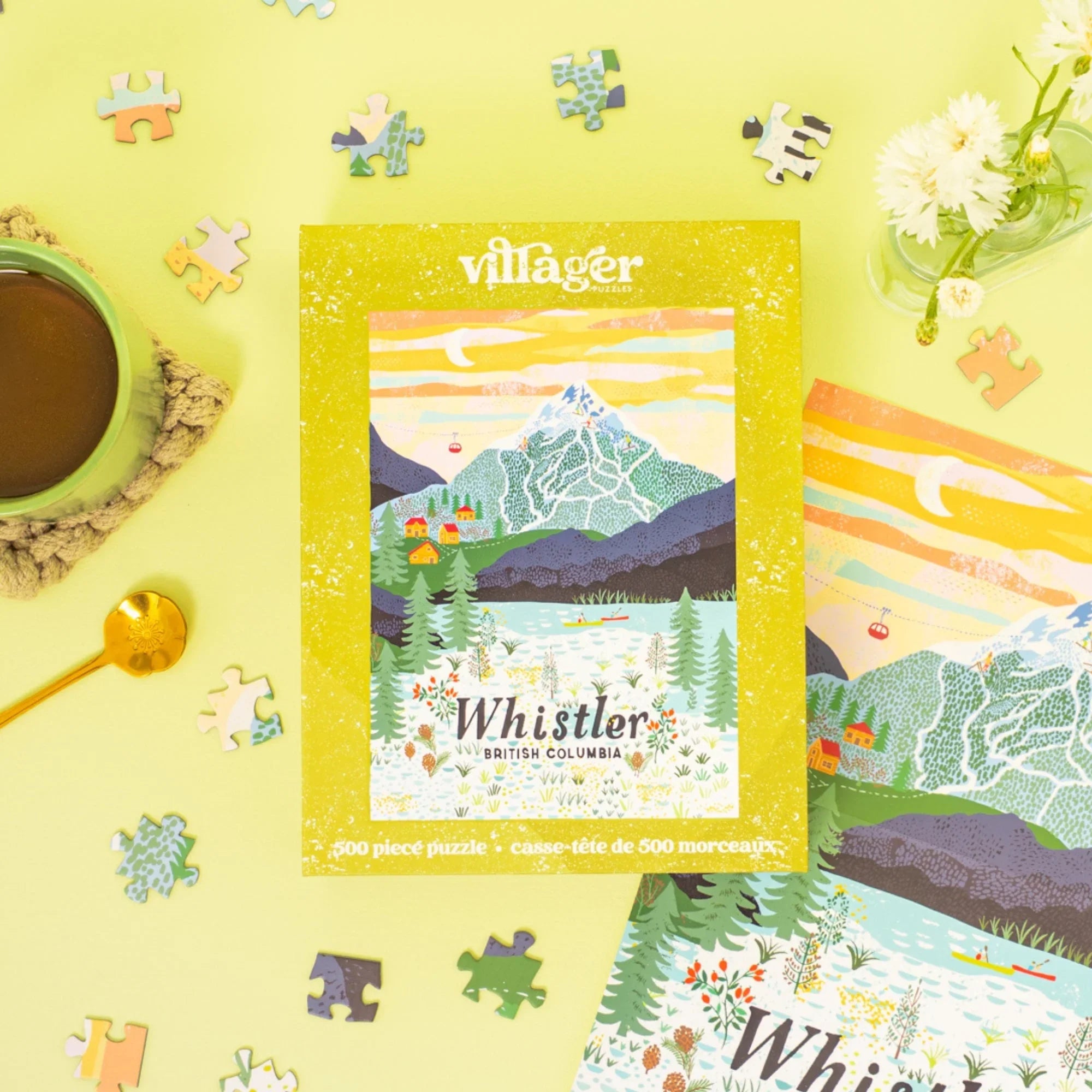 Whistler British Columbia jigsaw puzzle box with mountain illustration, coffee, flowers, and puzzle pieces on a yellow surface.