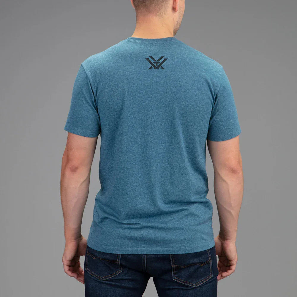 Man in blue crew neck t-shirt with black logo on back, dark jeans, gray background