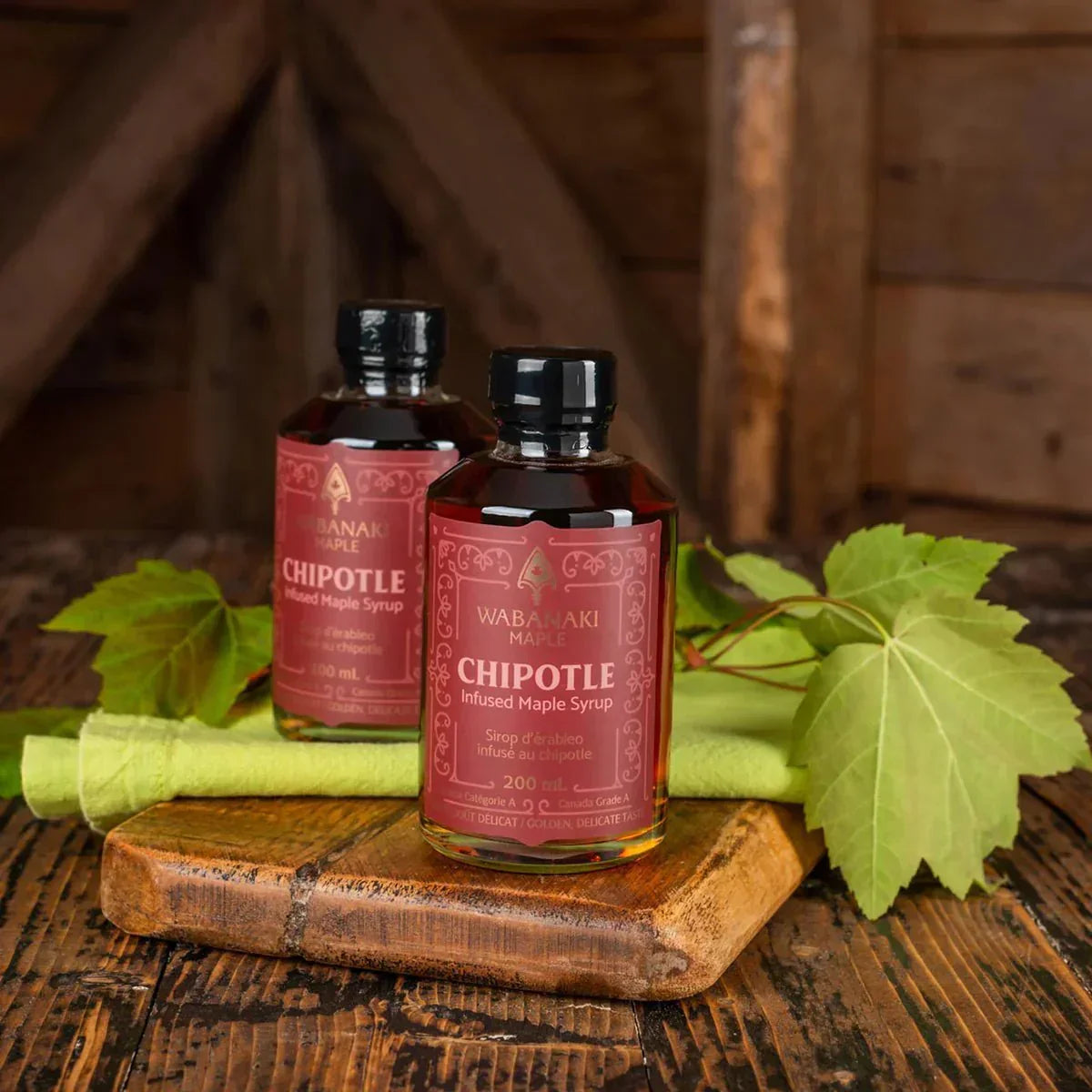 Wabanaki Maple Chipotle infused maple syrup bottles with green maple leaves on rustic wood.