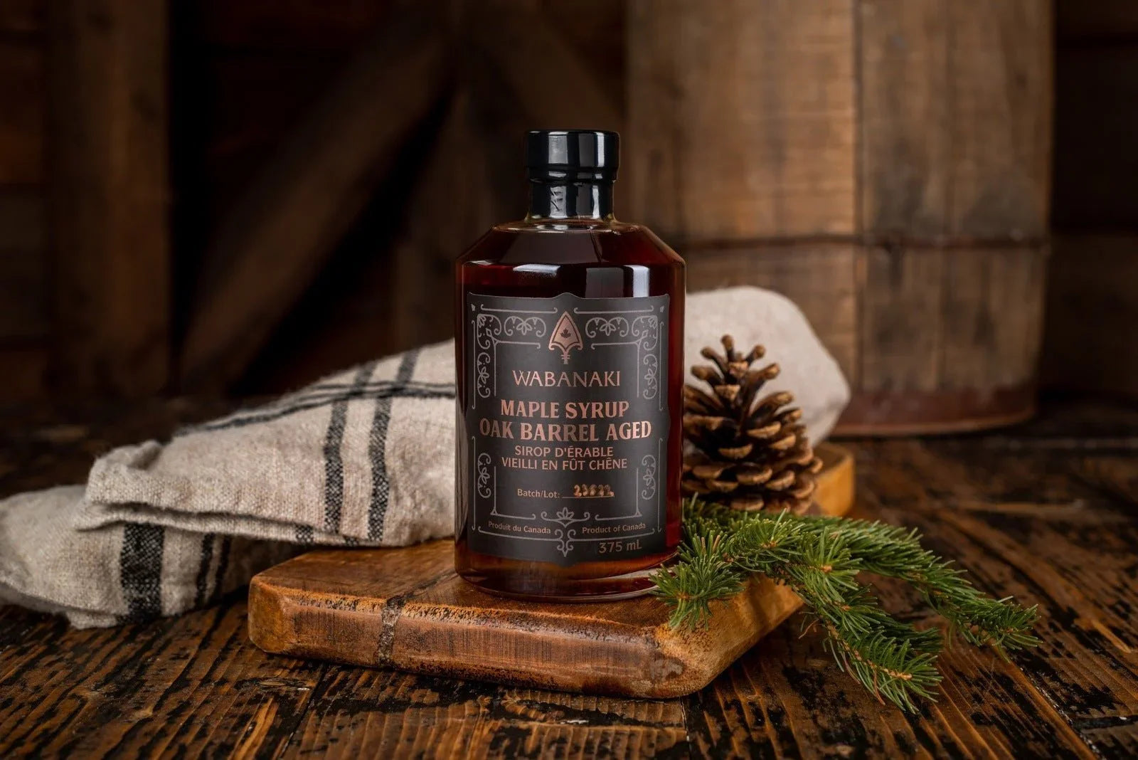 Wabanaki Maple Syrup oak barrel aged bottle on rustic wood with pinecone and evergreen sprig