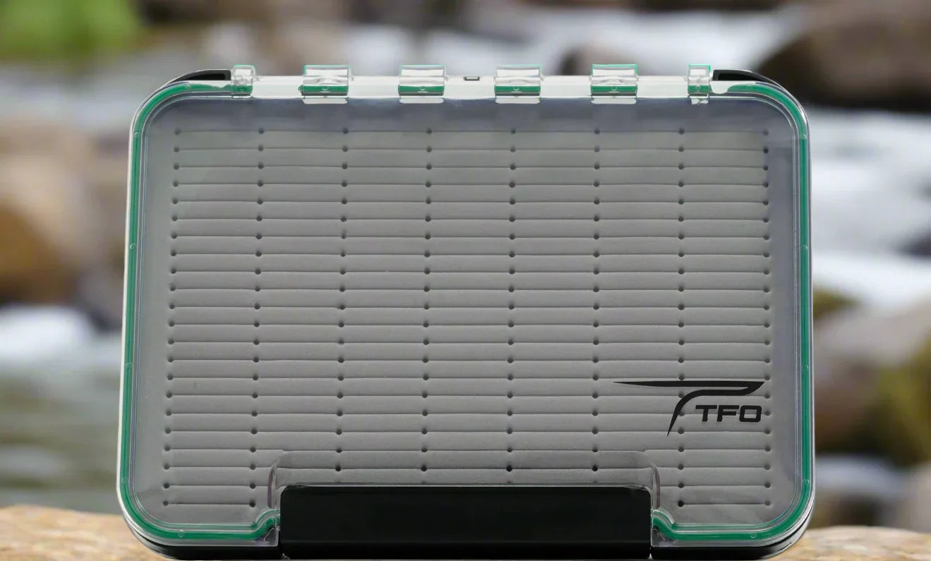 TFO waterproof fly box with clear lid and foam insert, outdoors on a rock by a stream