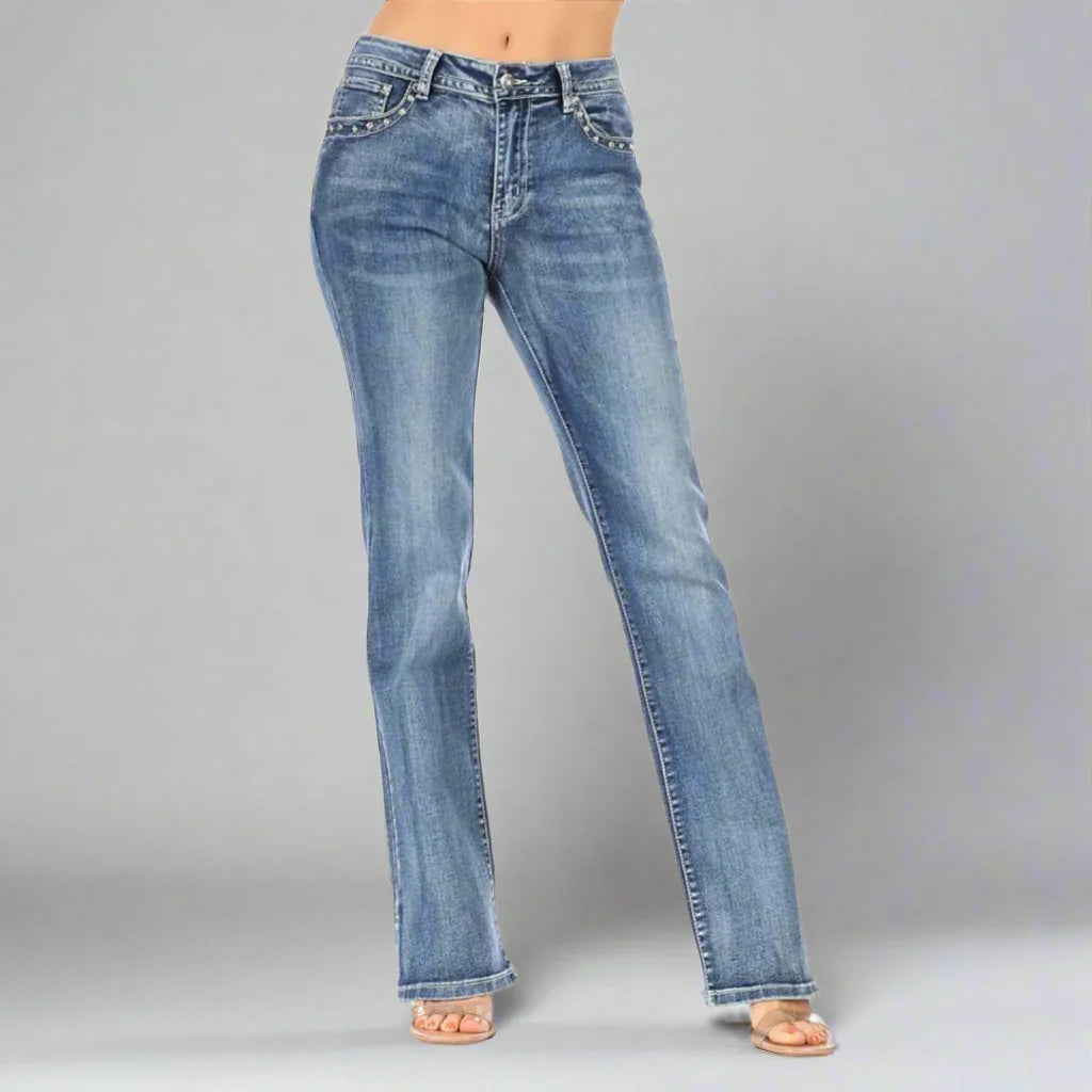 Woman modeling medium wash bootcut jeans with studded pockets on a gray background