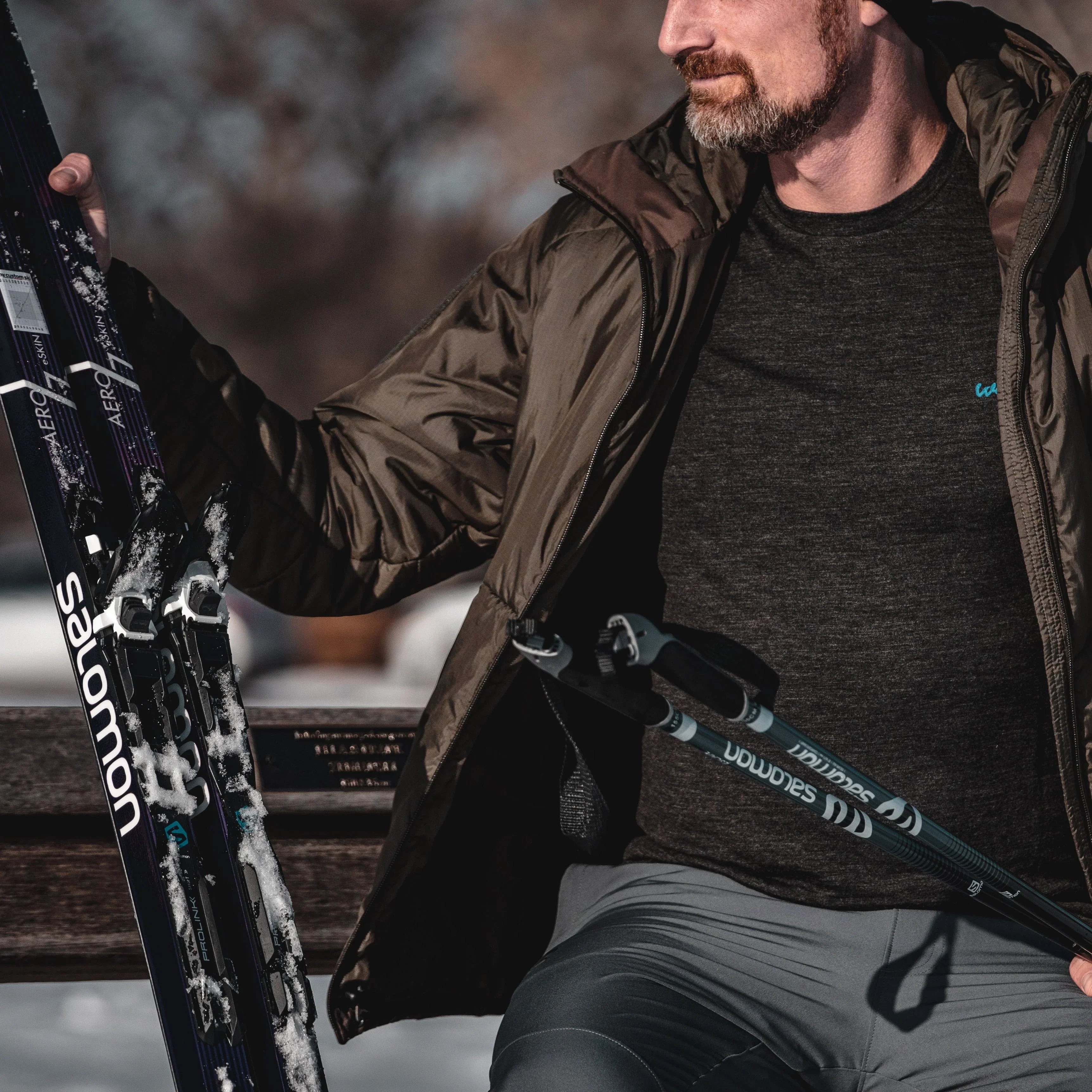 Man in outdoor winter clothing holding Salomon skis and poles on a snowy bench