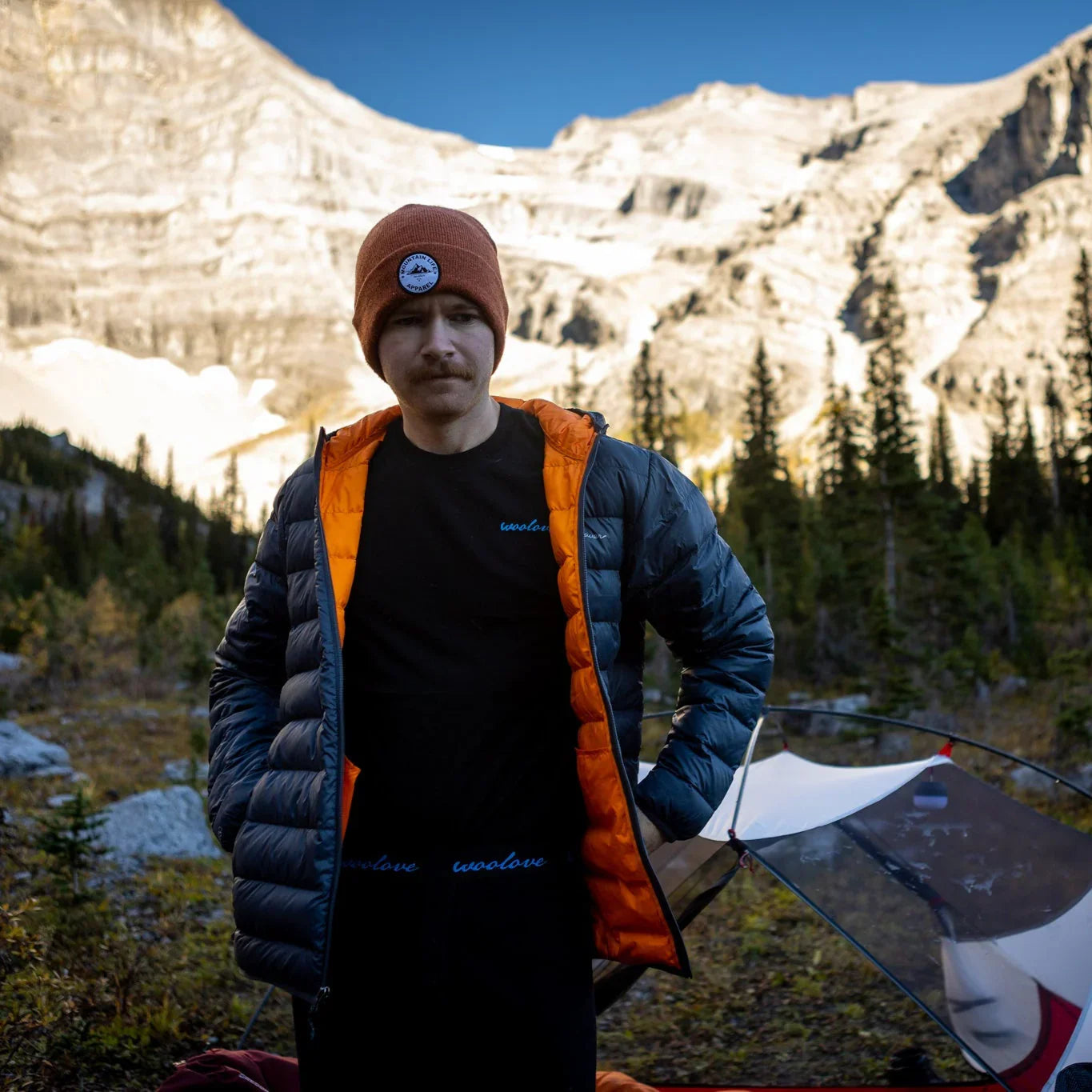 Man in Woolove apparel puffer jacket and beanie standing by tent in mountain landscape