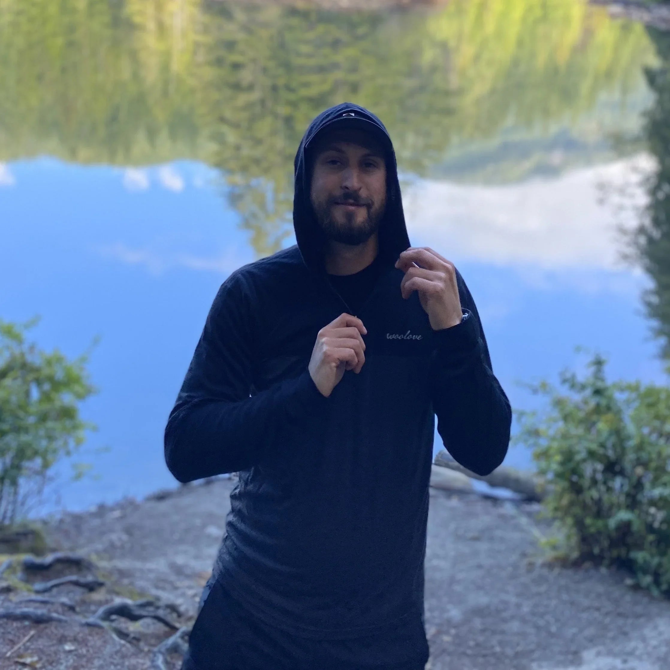 Man wearing a black quarter zip hoodie outdoors by a lake with trees reflected in water