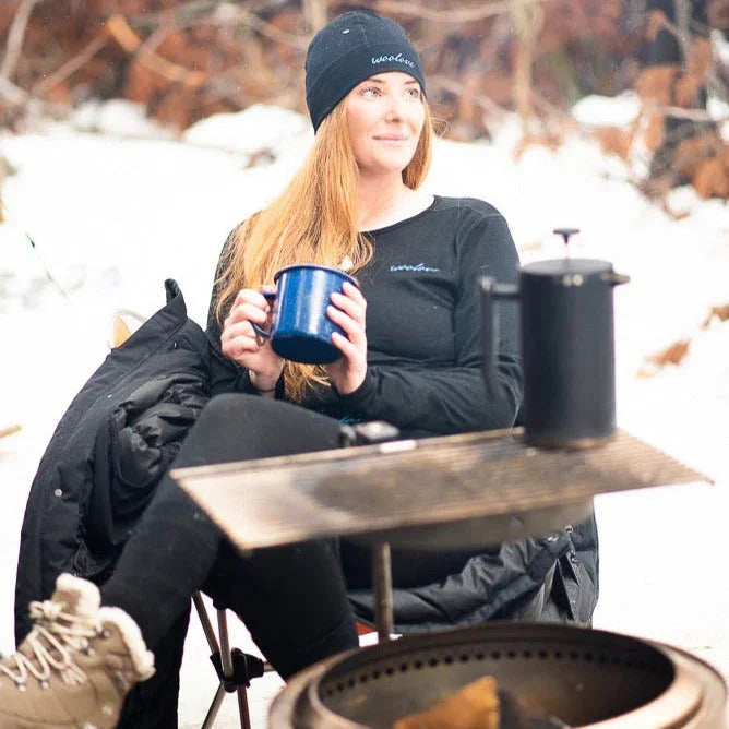 Woman in black beanie and long sleeve shirt camping in the snow, holding blue mug by campfire.