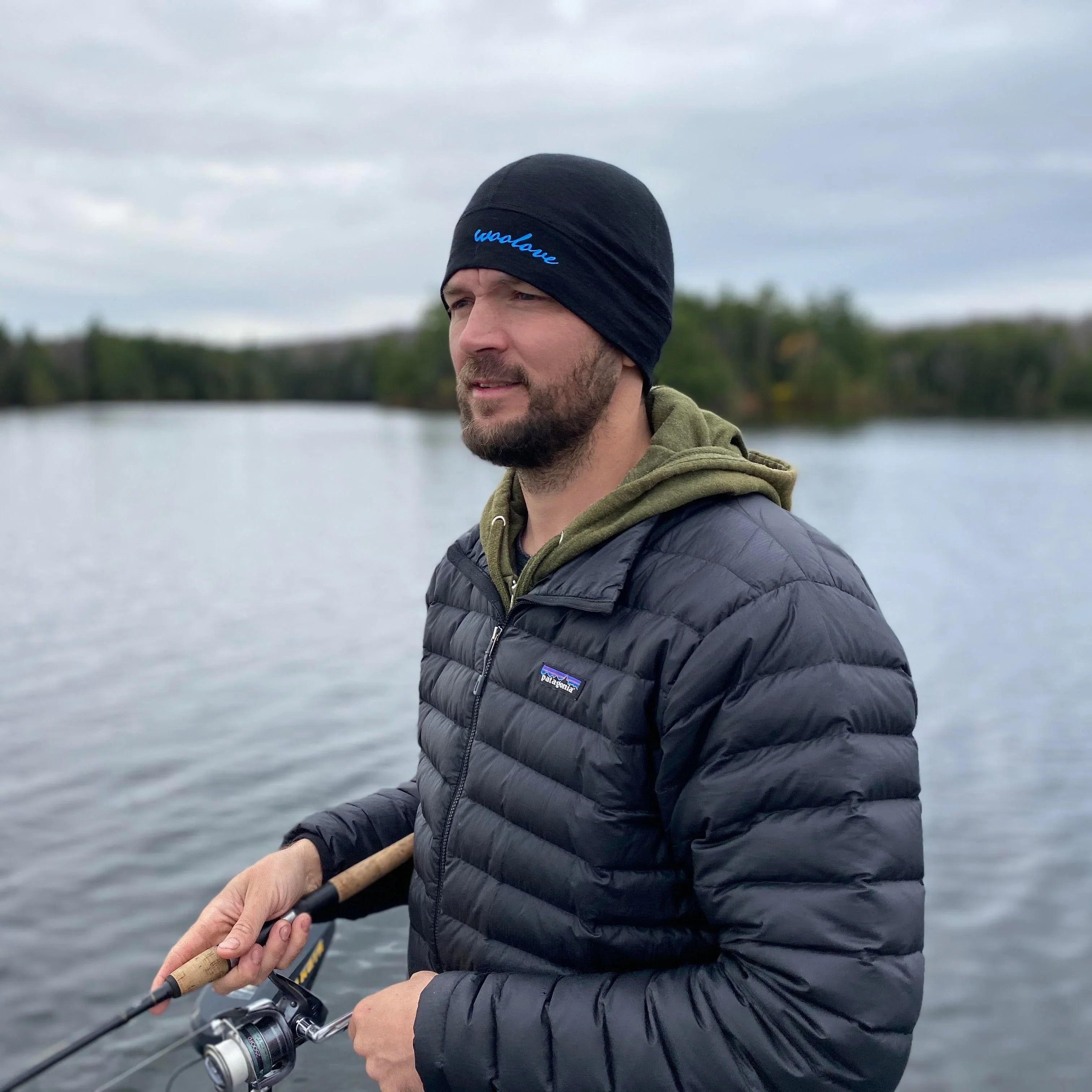 Man fishing outdoors wearing black 100% merino wool sport beanie and puffer jacket by lake