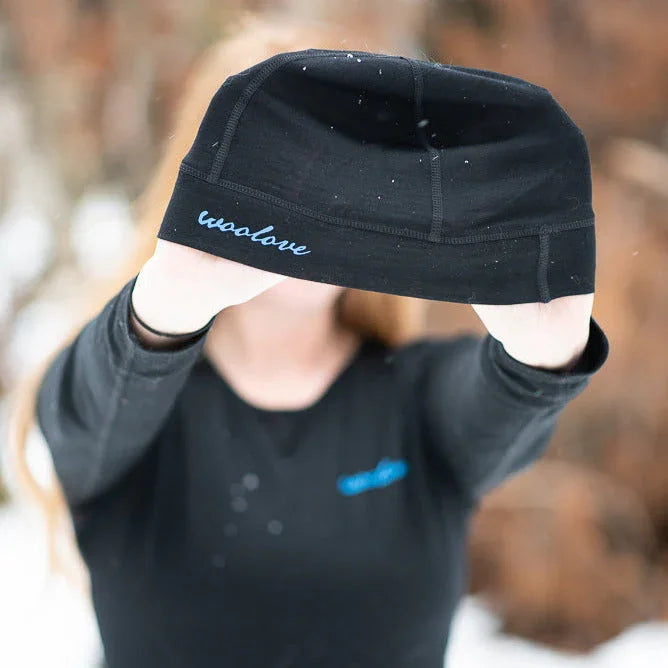 Woman holding black Woolove beanie outdoors in snow, winter clothing product photo