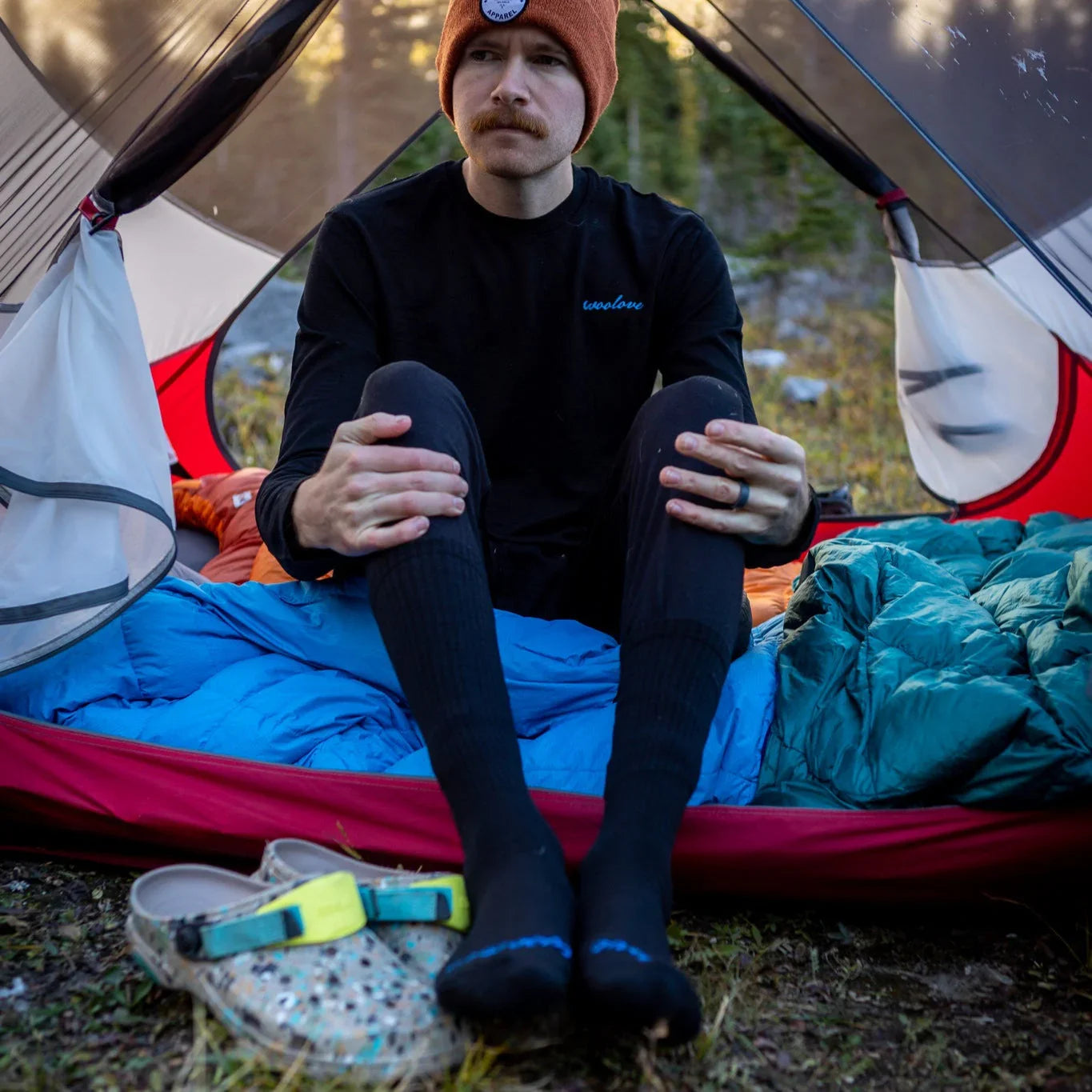 Man in black Woolove base layer sitting in tent with sleeping bag and camping gear outdoors