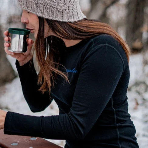 Woman in knit beanie and black long sleeve shirt drinking from metal mug outdoors in winter
