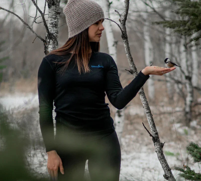 Woman in black long sleeve outdoor top feeds bird in snowy forest, wearing knit beanie