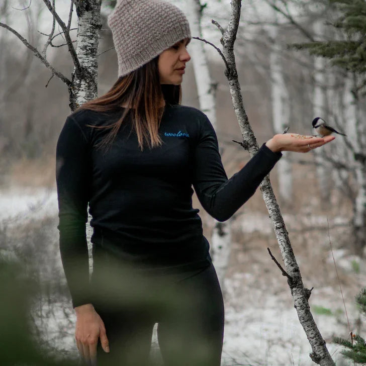 Woman in black Woolara top and knit hat holding bird outdoors in snowy birch forest