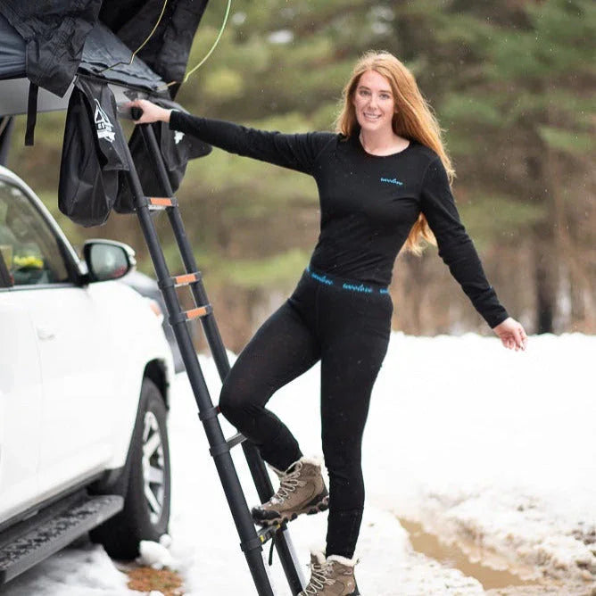 Woman in black thermal base layers climbing a rooftop tent ladder beside SUV in snowy outdoors