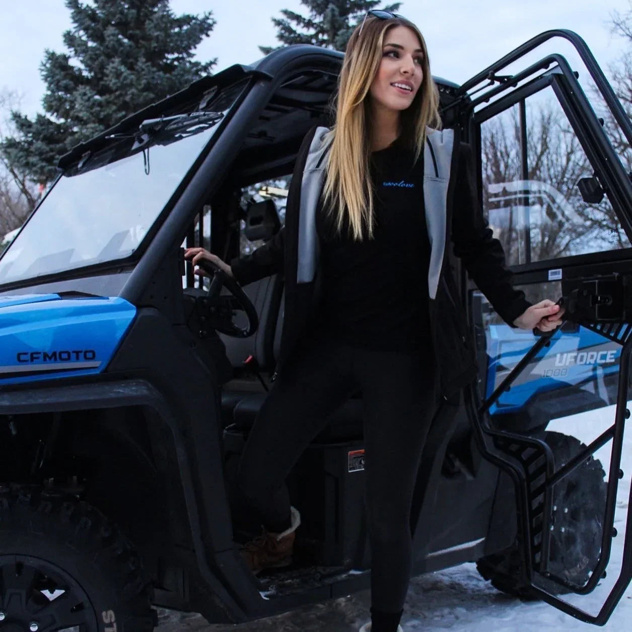 Woman in black leggings and jacket stepping out of a blue CFMOTO UTV in snowy outdoor setting
