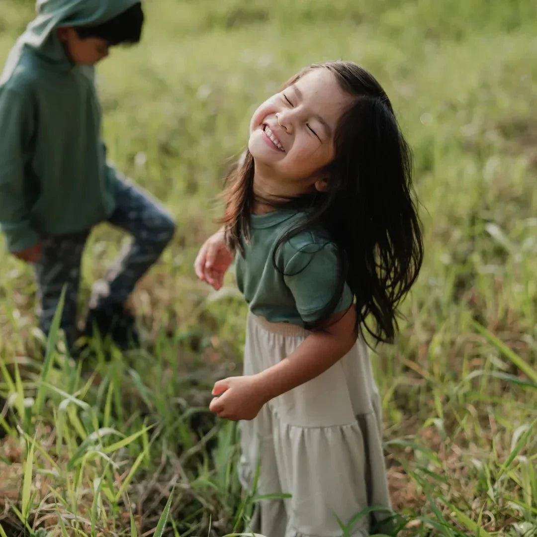 Smiling child in a bamboo aria skirt and green top standing in grassy field outdoors
