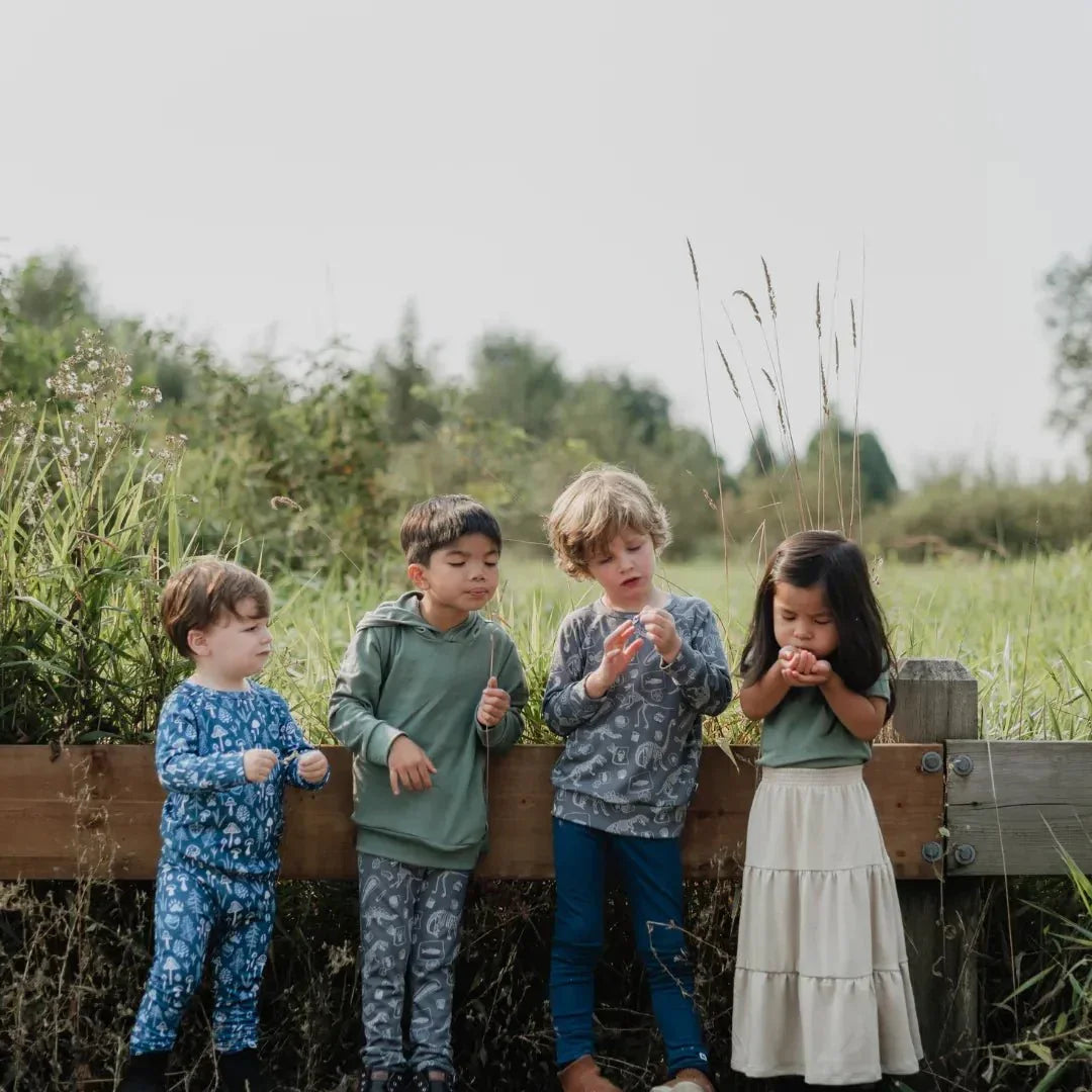 Four kids in bamboo clothing stand outdoors by a wooden fence in a grassy field