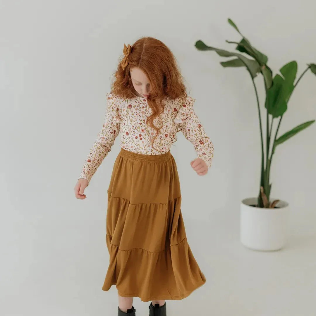 Girl in brown bamboo Aria skirt and floral top stands indoors near potted plant