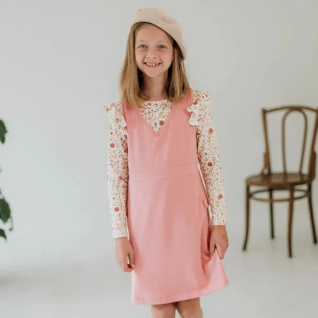 Girl wearing pink bamboo cargo jumper dress and floral shirt in a minimal room