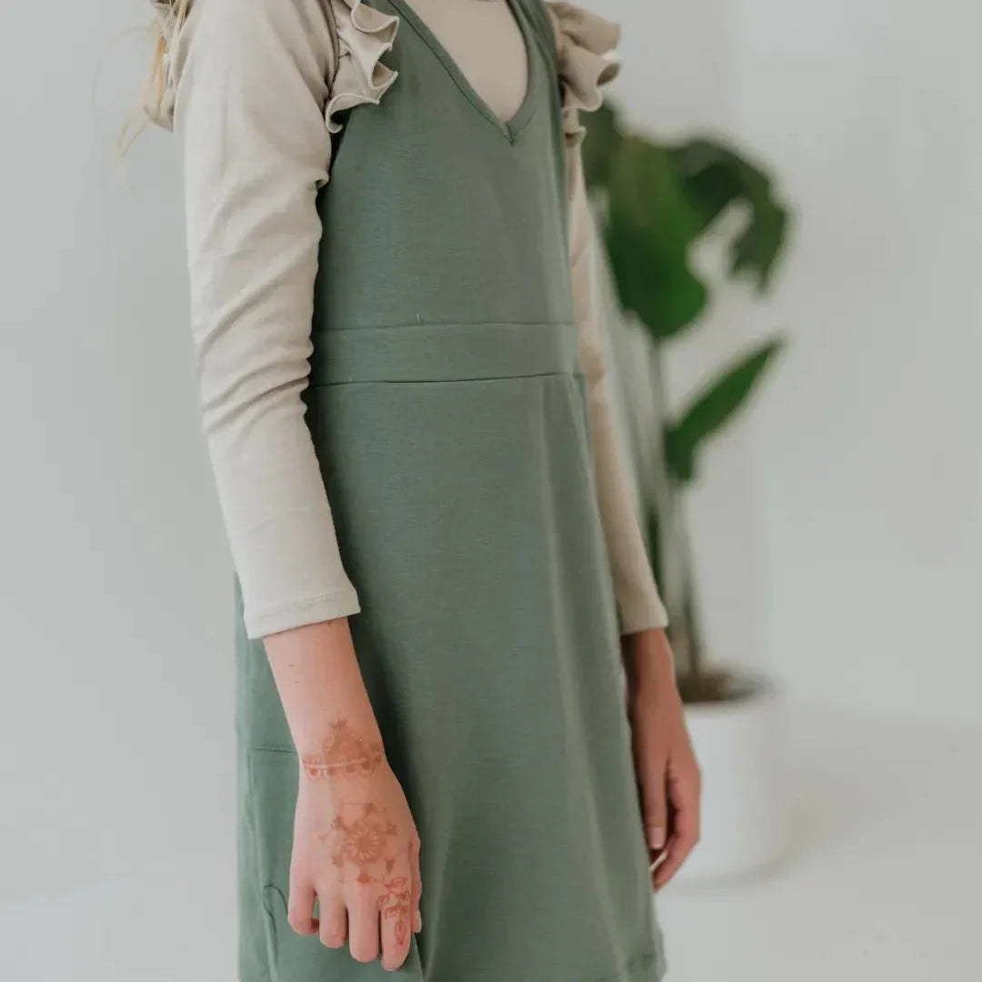 Girl wearing green bamboo cargo jumper dress over ruffle long-sleeve shirt, plant background