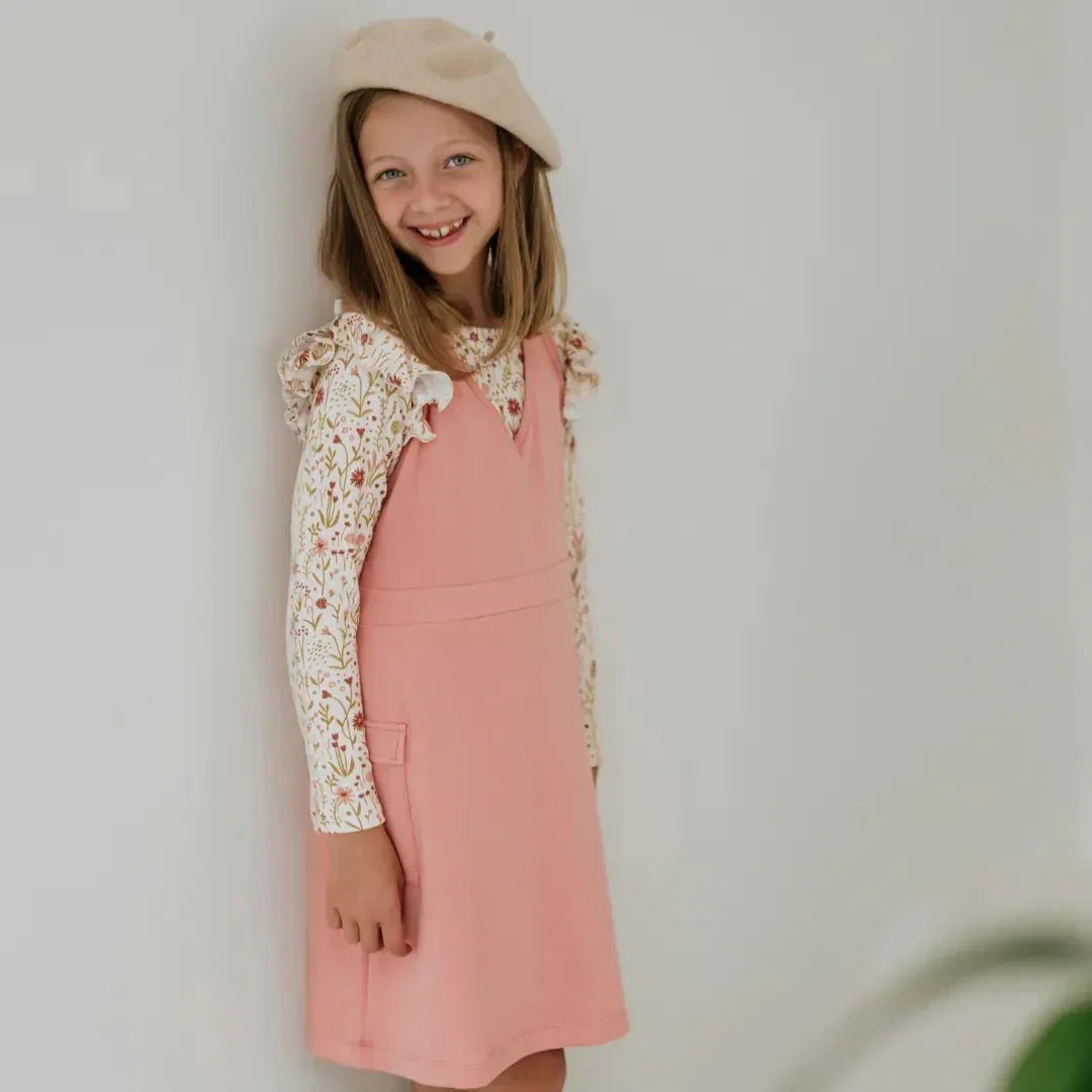 Smiling girl in a pink bamboo cargo jumper dress and floral top, wearing a beige beret