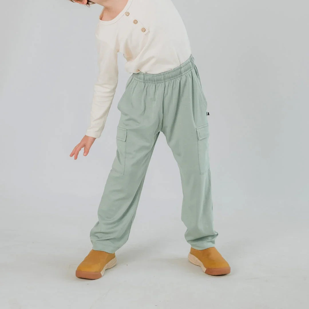 Child in sage green cargo pants and cream long-sleeve top with tan shoes on white background