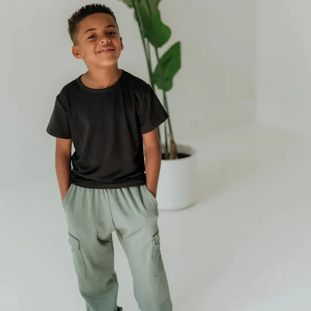 Smiling boy in black t-shirt and sage green bamboo cargo pants in minimal white room