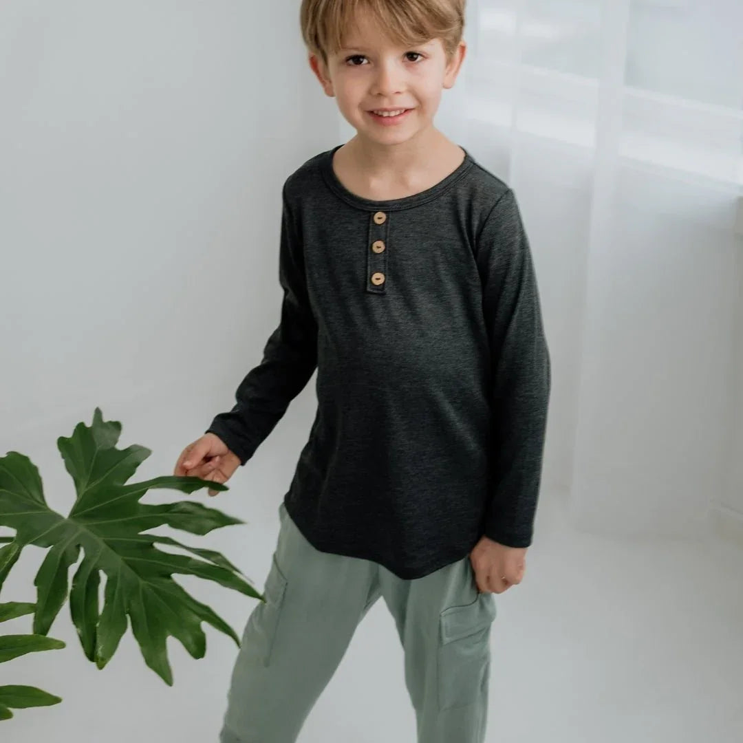 Smiling boy in dark bamboo Henley long sleeve shirt and light pants indoors by green plant