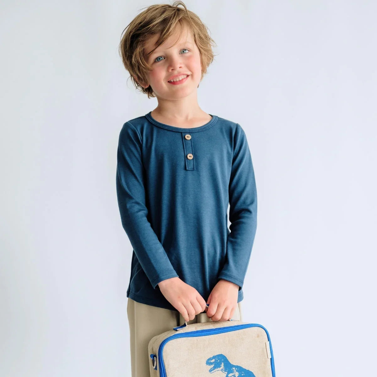 Smiling child in blue long sleeve shirt holding dinosaur lunch bag against white background