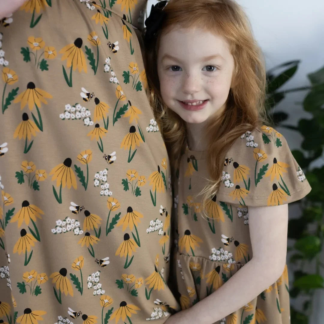 Smiling girl in matching yellow floral dress with bees, standing beside adult in same print