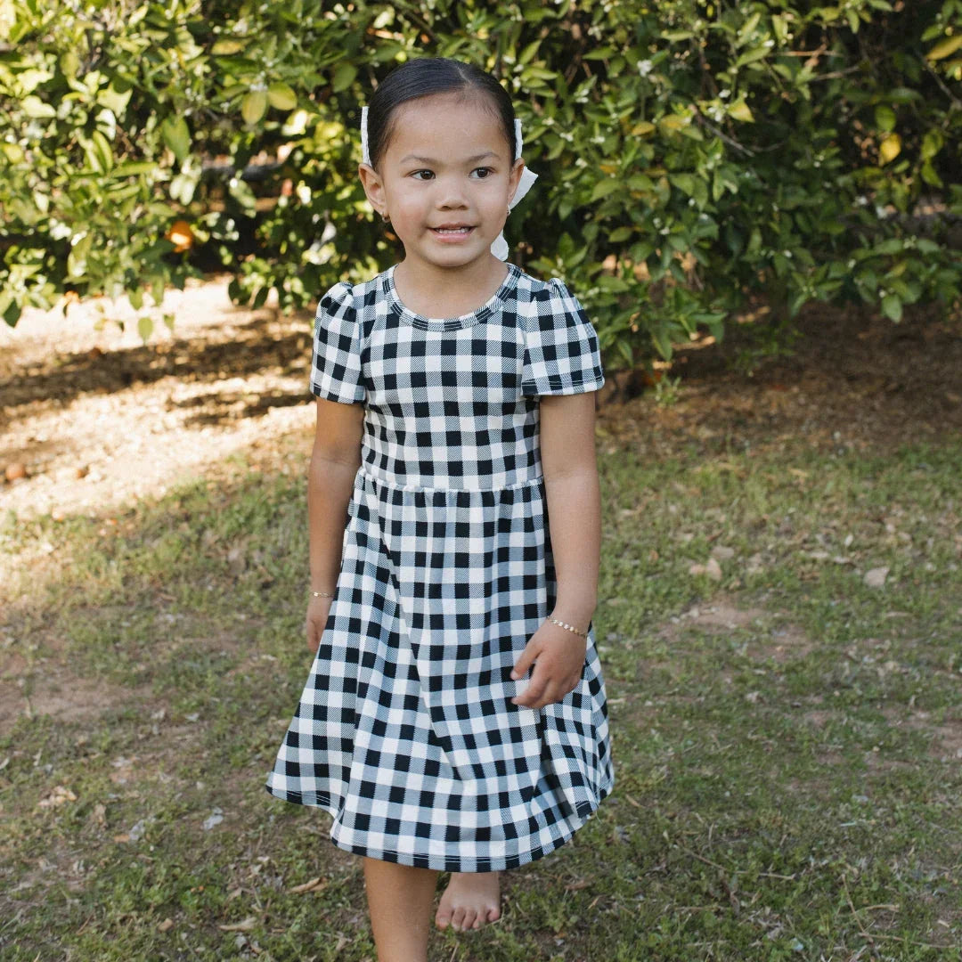 Girl in black and white gingham dress standing barefoot outdoors near green bushes