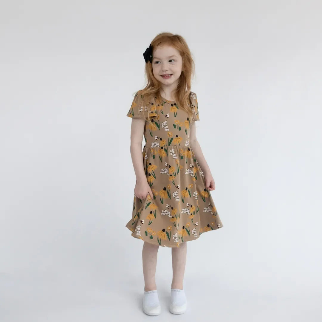 Girl in floral print brown dress and white shoes standing against plain white background
