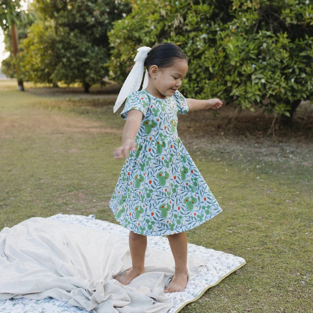 Smiling girl in floral dress with white bow, spinning barefoot on blanket outdoors in garden