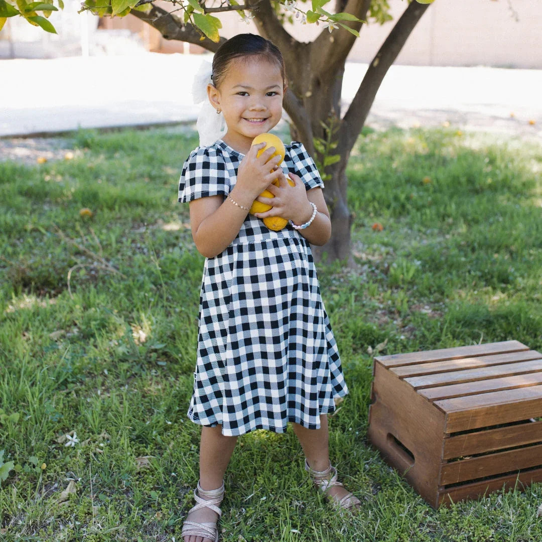 Smiling girl in a black and white plaid dress holding lemons under a tree in a garden