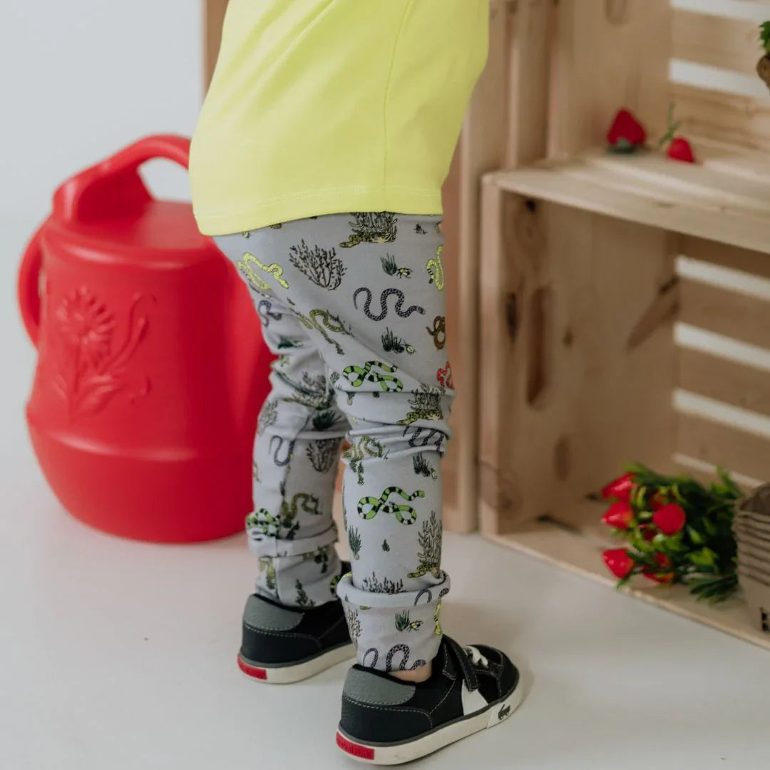 Child in snake print leggings and yellow shirt near red watering can and wooden crate