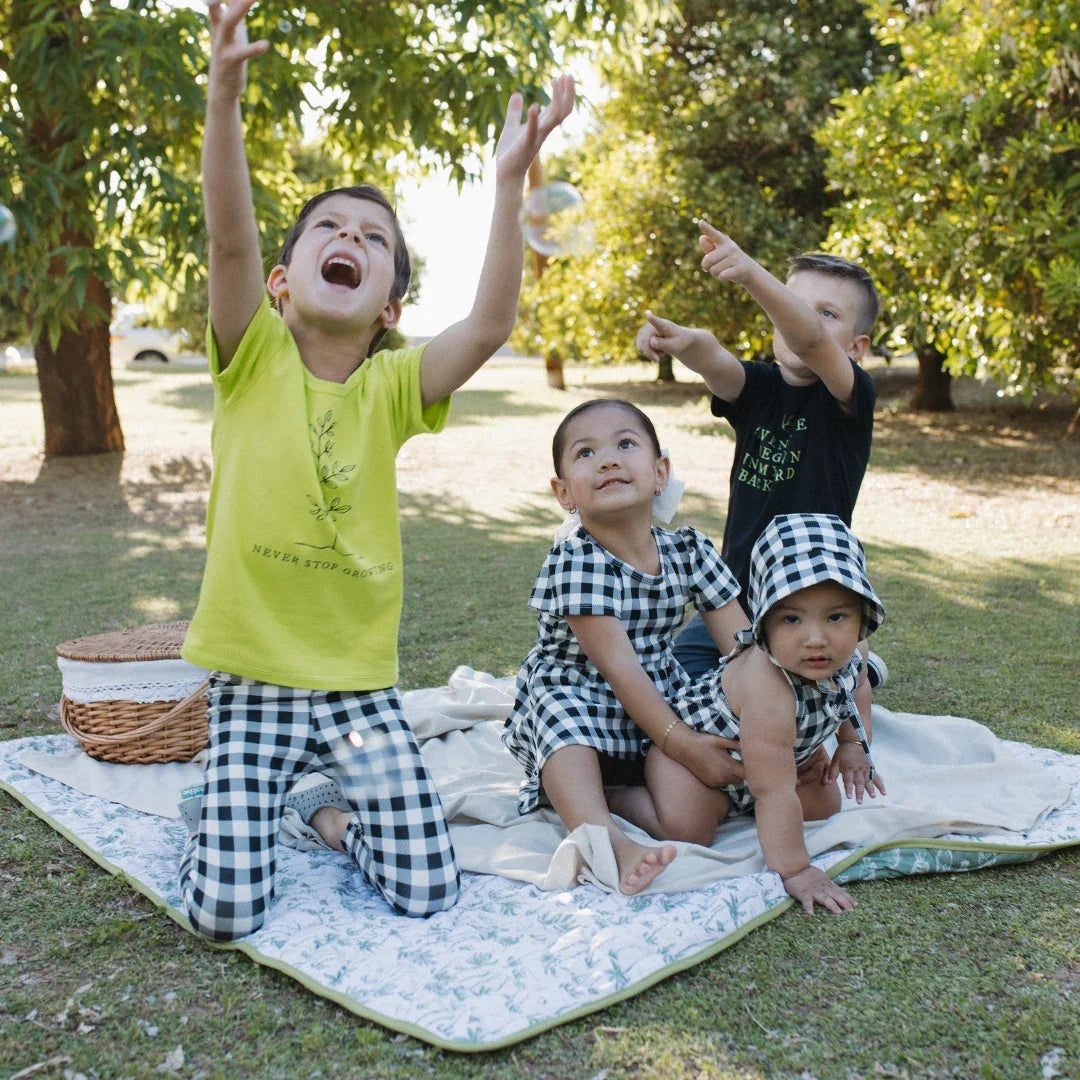 Four kids in checkered outfits and green shirts play outdoors on a picnic blanket under trees.