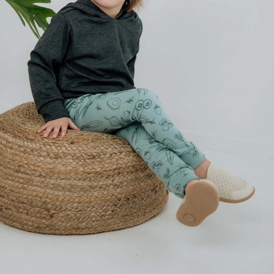 Toddler in bamboo print leggings and dark hoodie sitting on woven pouf, indoor setting