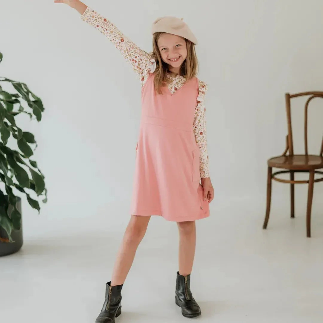 Smiling girl in a pink dress and beret posing in a bright room with a wooden chair and plant