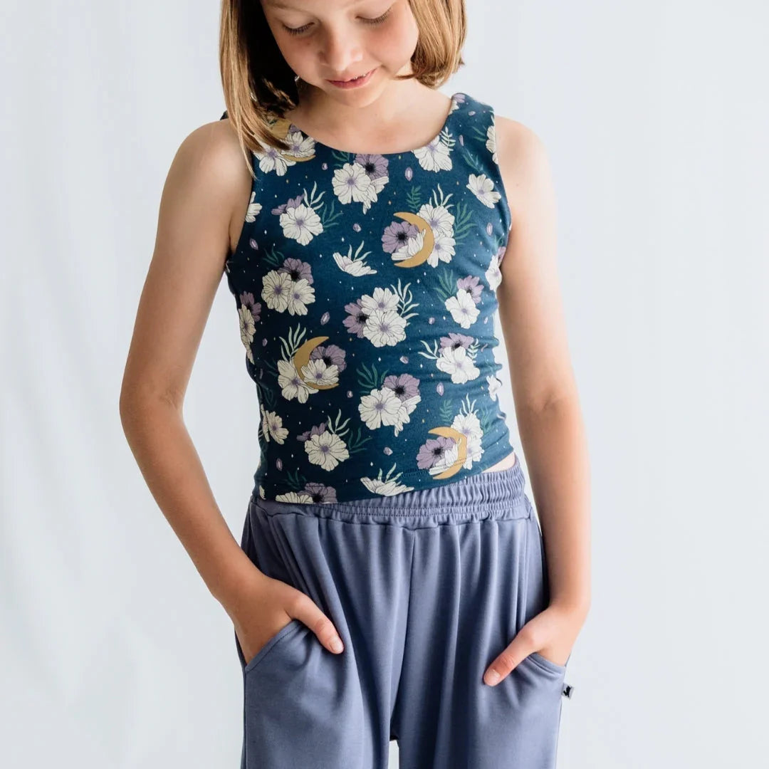 Girl wearing floral tank top with moon print and blue pants, hands in pockets, white background