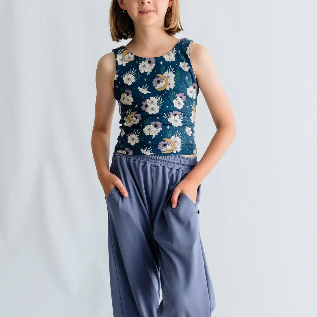 Girl wearing floral tank top and loose blue pants, hands in pockets, plain background