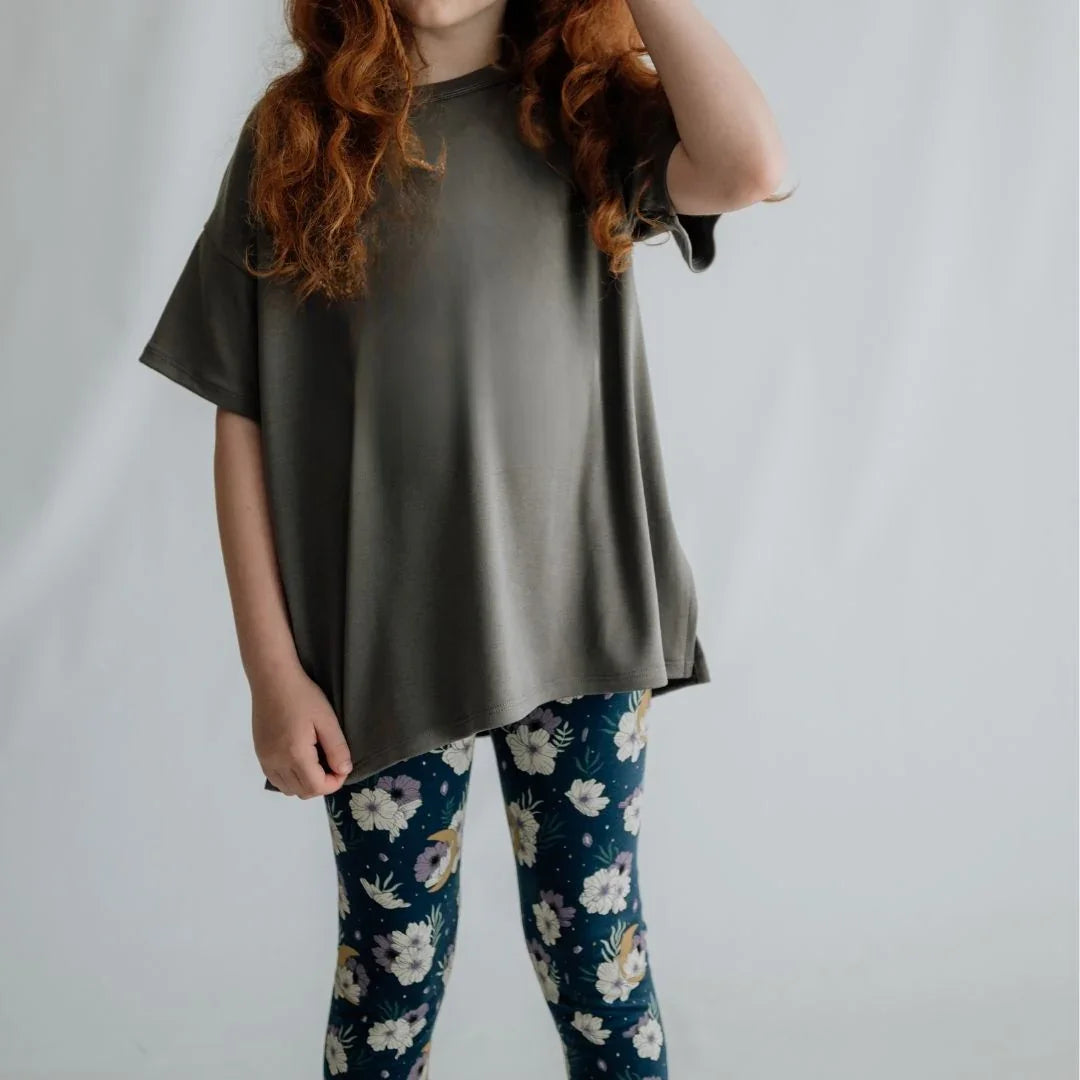 Child with long red hair wearing loose gray shirt and floral print leggings, studio background
