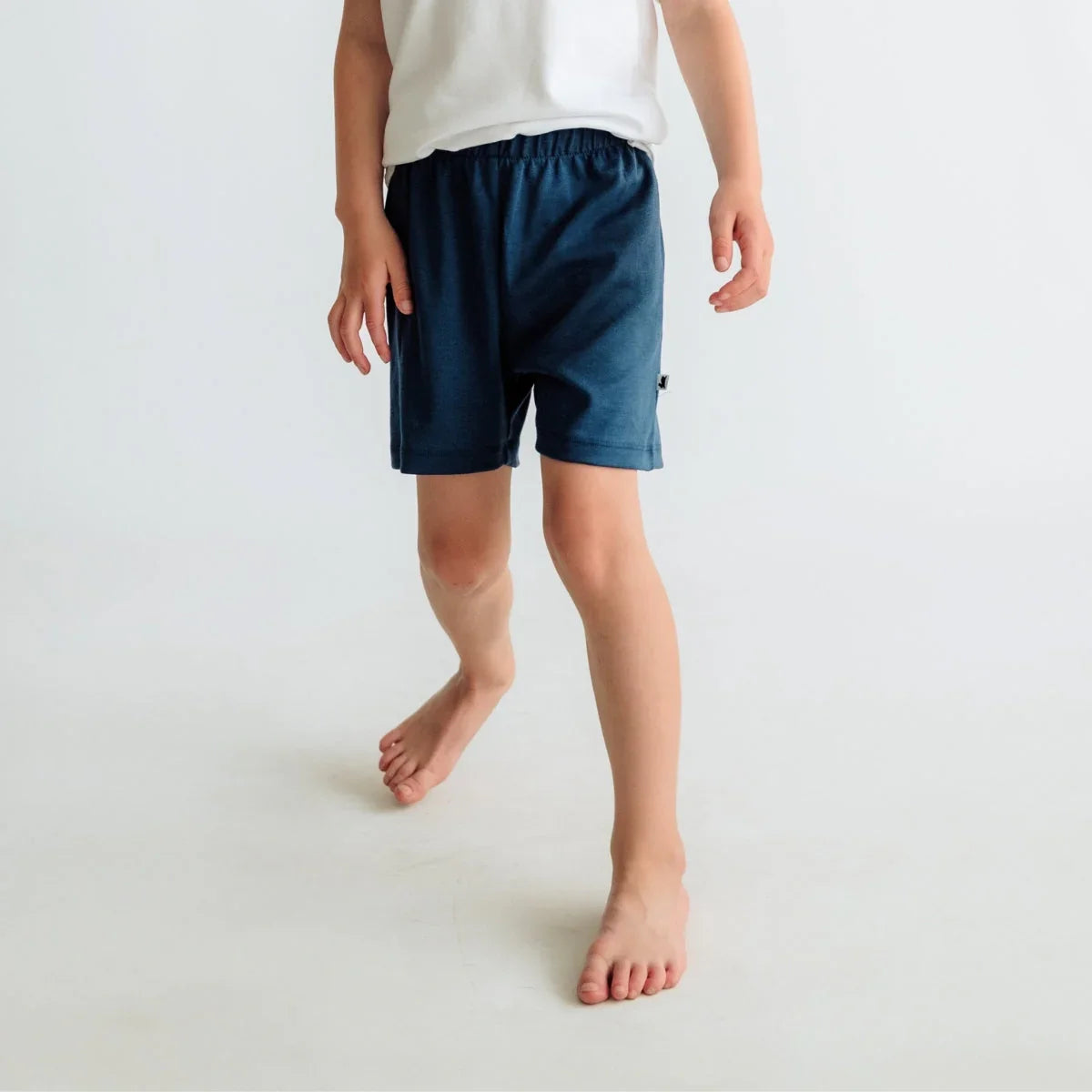 Child in white shirt and navy blue shorts standing barefoot on a plain light background