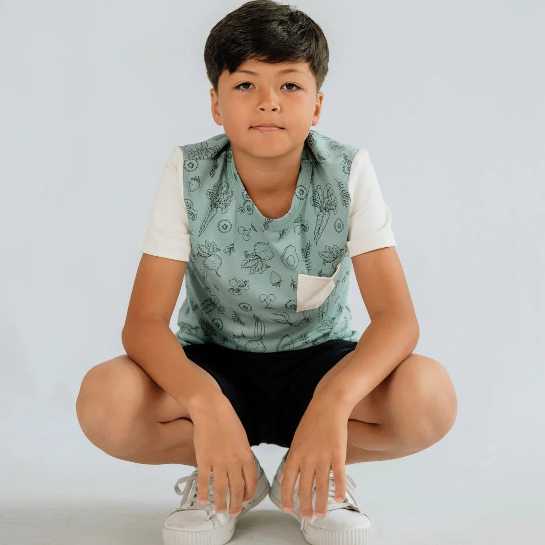 Youth boy wearing mint green pocket tee with fruit print, white sleeves, squatting indoors